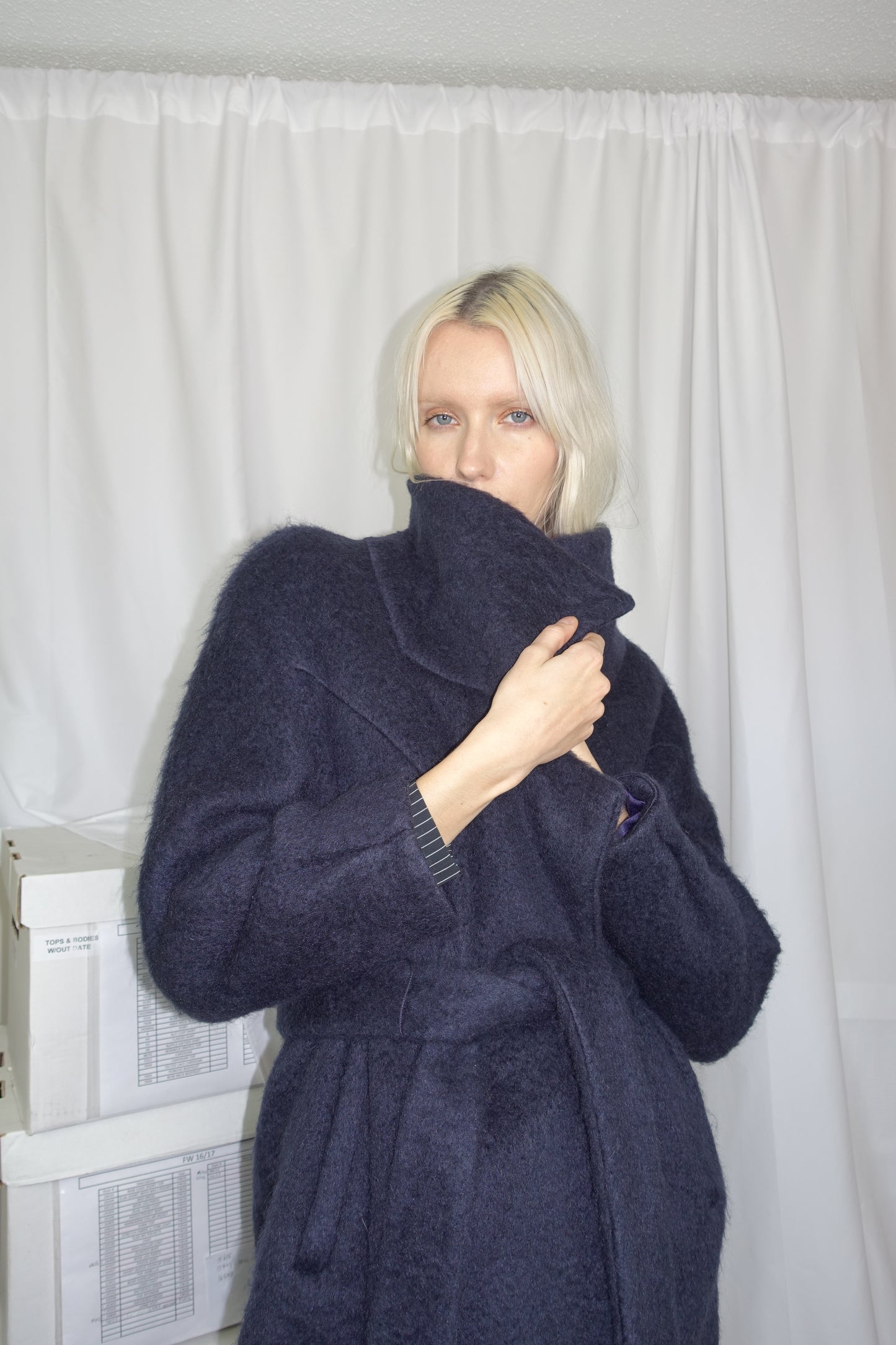 Woman wearing a midnight colored mohair double breasted coat with wide collar cut and double self tie belt. Concealing her mouth ominously.