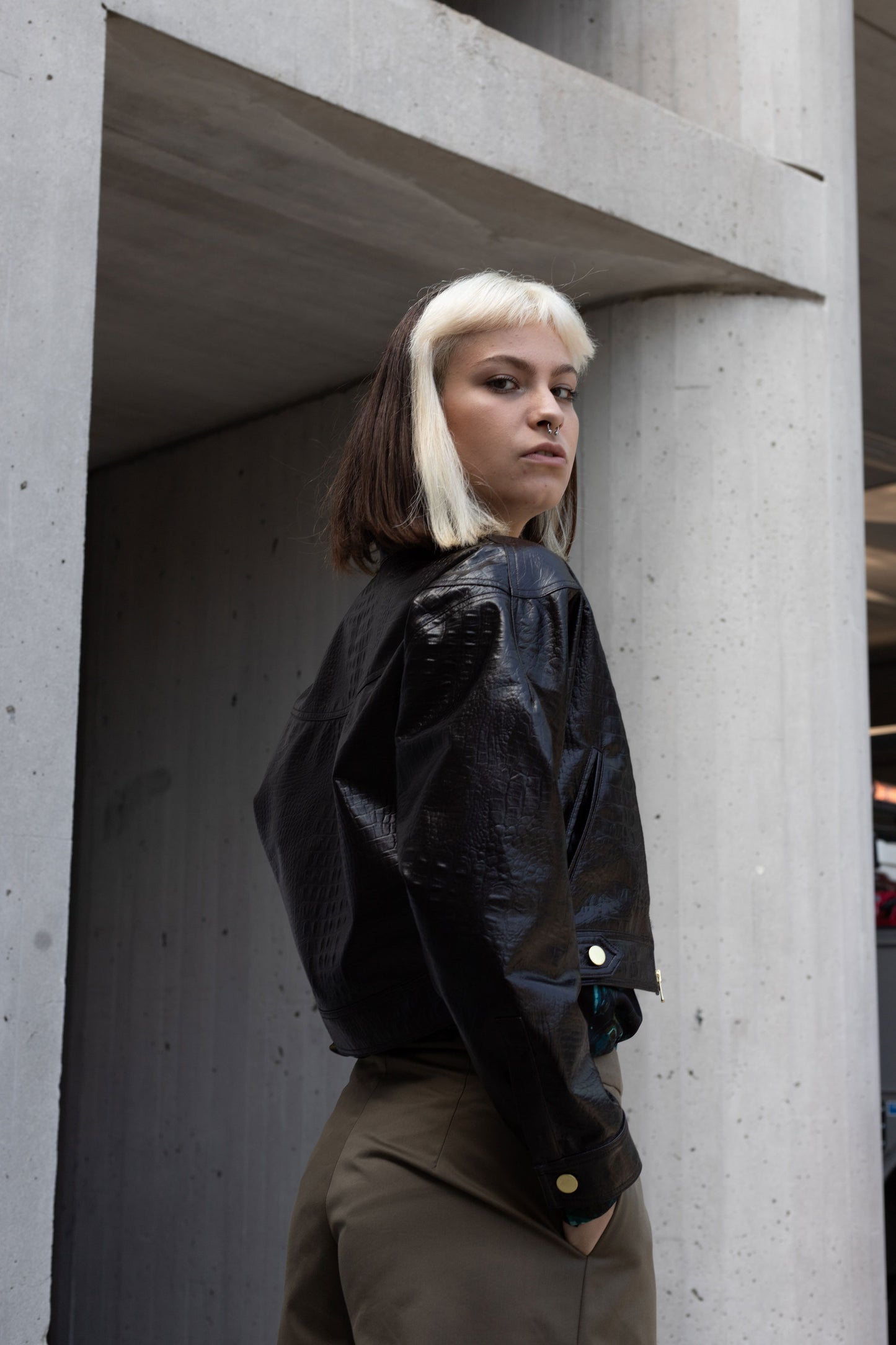 Back of woman wearing a chocolate brown cropped waist jacket, with embossed and coated crocodile pattern and modern silhouette with detailed seaming over gray pants