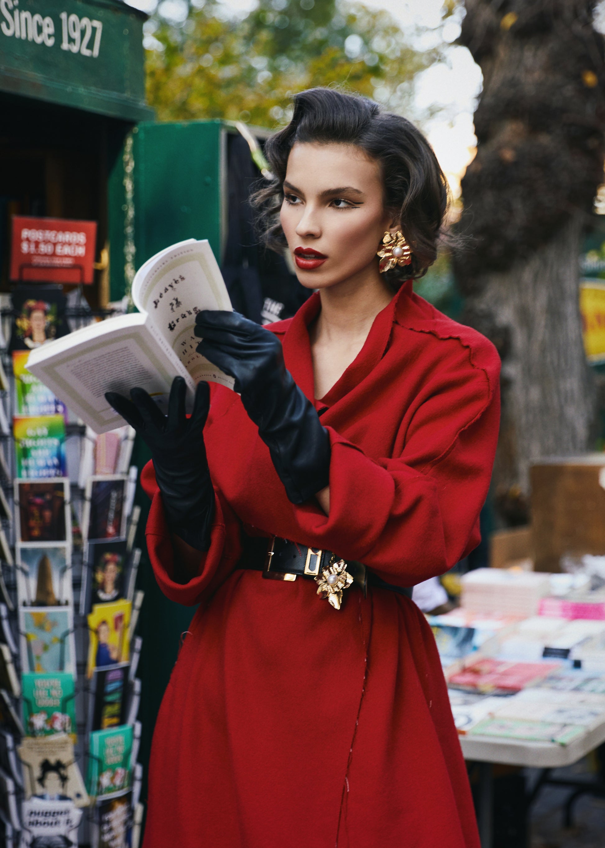 A woman wearing a red rumi coat with a black-golden accented waist belt. She is reading a book in an outdoor bookstand.

By @monicabaddar
Featuring our Rumi coat
Modeled by @savka.dm
Styled by @styledbybruce
Hair by @michikoboorberg
Make up @brennadmakeup
Courtesy @the_confessional_showroom_nyc