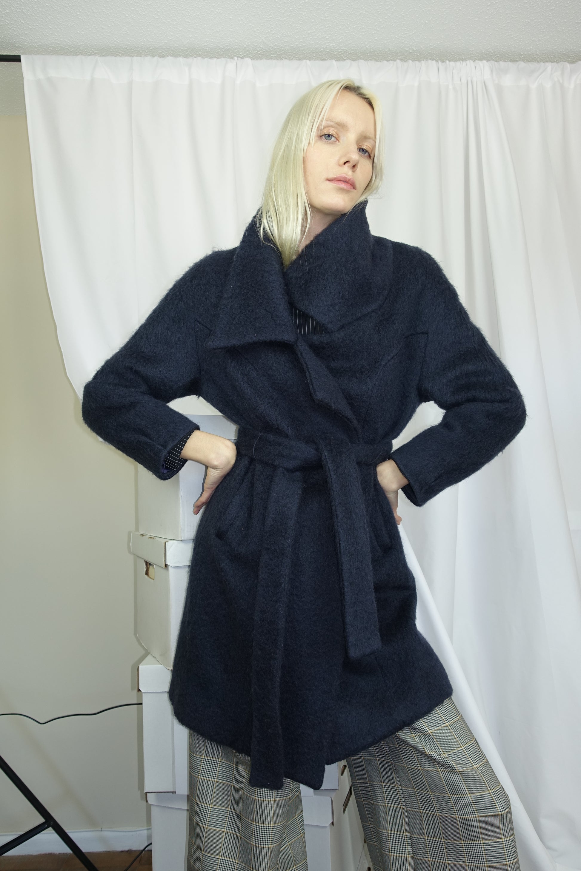 Woman wearing a midnight colored mohair double breasted coat with wide collar cut and double self tie belt. Posing in a studio room with hands on hips.