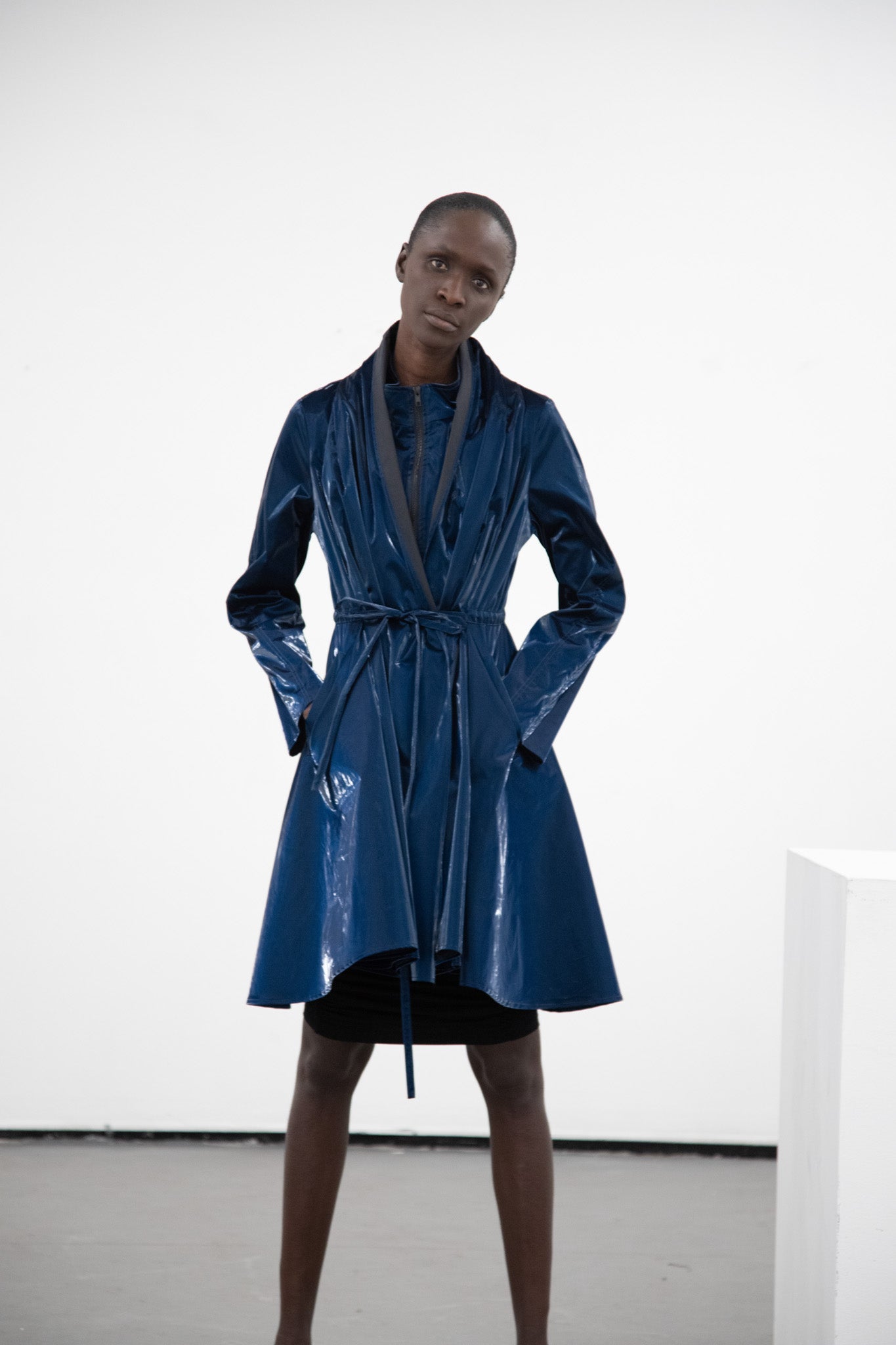 Women posing with hands in pocket and a titled head wearing a blue coat featuring wrinkle fabric combined with a tie-belt.