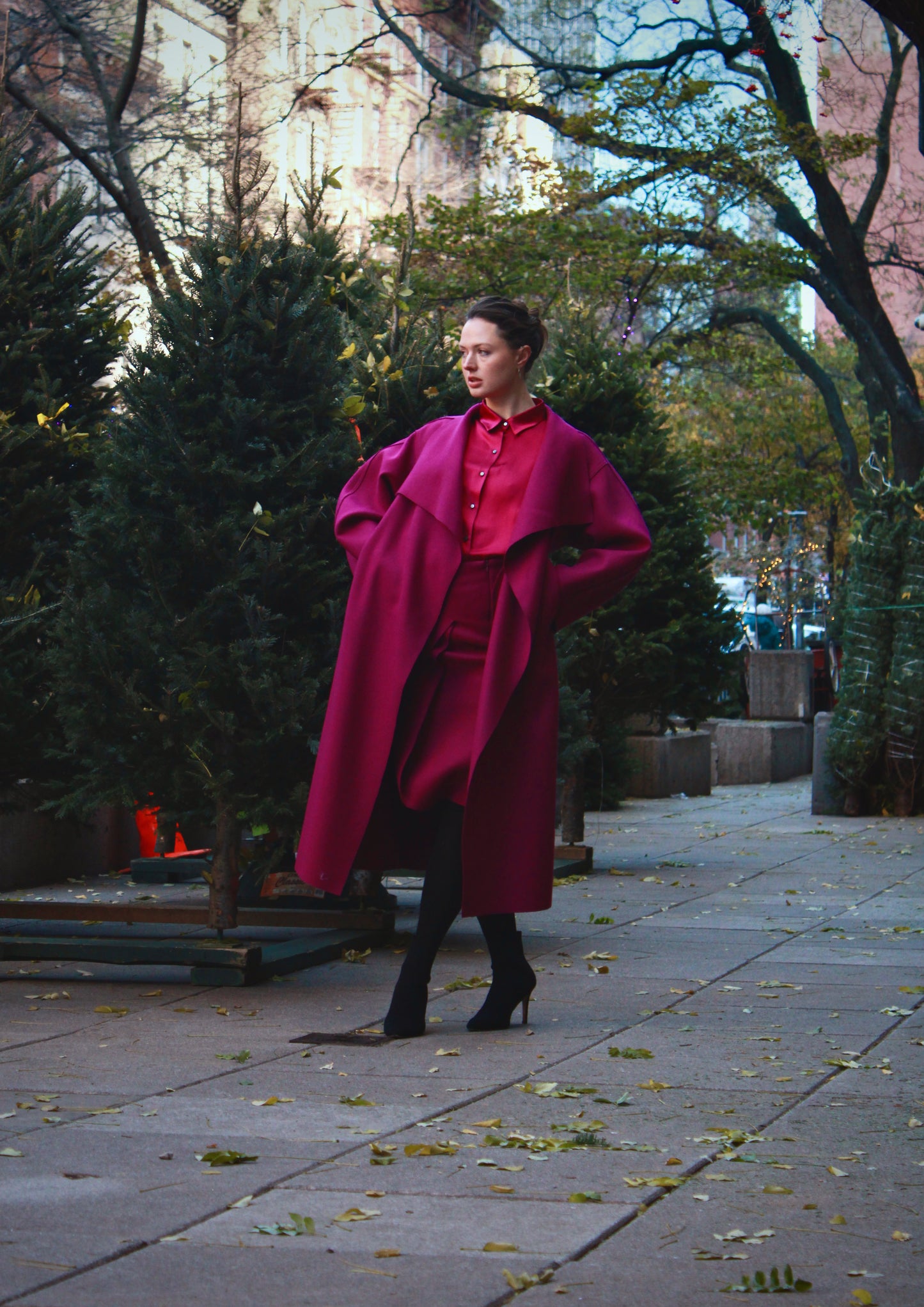 Woman standing in an outdoor scene with greenery in the background wearing a purple rumi coat with a red collared shirt and black high heels.