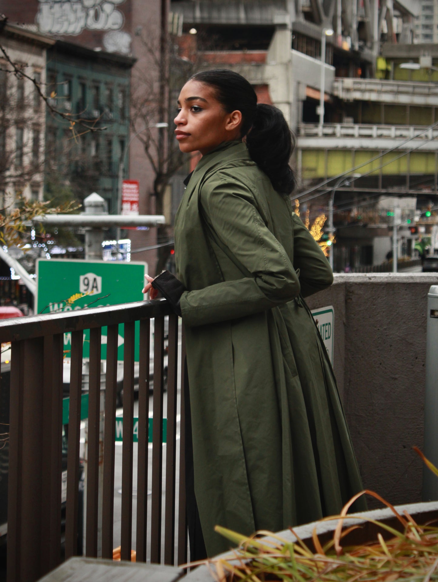 Woman posing on an outside balcony fence wearing an olive Japanese nylon coat, looking at the street.