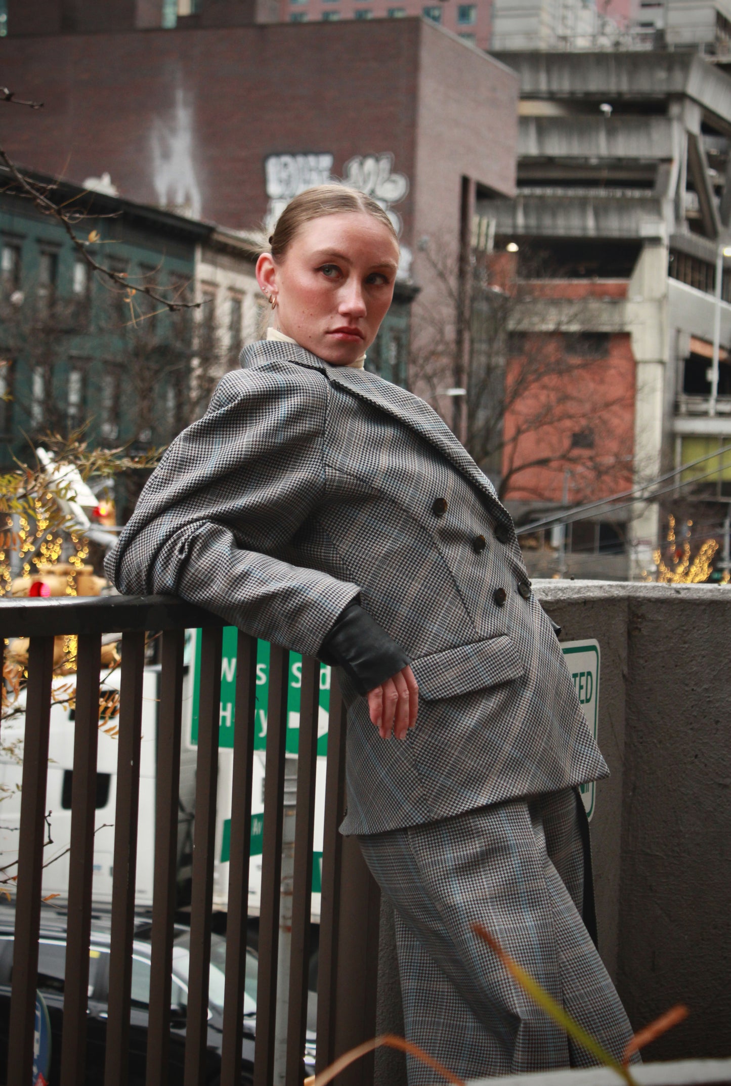 Woman wearing a gray double-breasted blazer with plaid patterns. Leaning casually on an outdoor street fence.