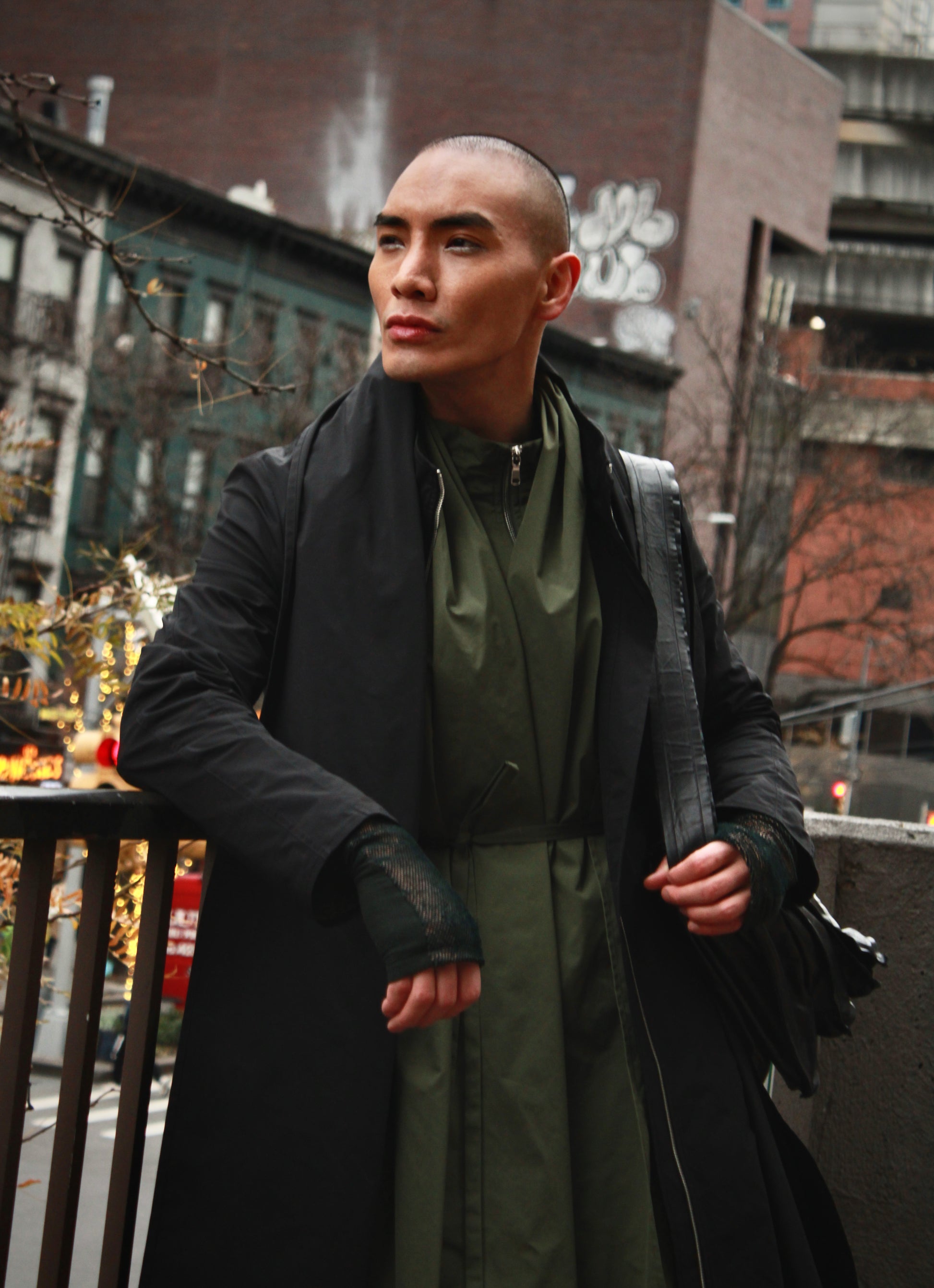 Person in an outdoor urban environment leaning on a balcony fence. Wearing black Japanese nylon raincoat and looking away.

Model: @lejamie_