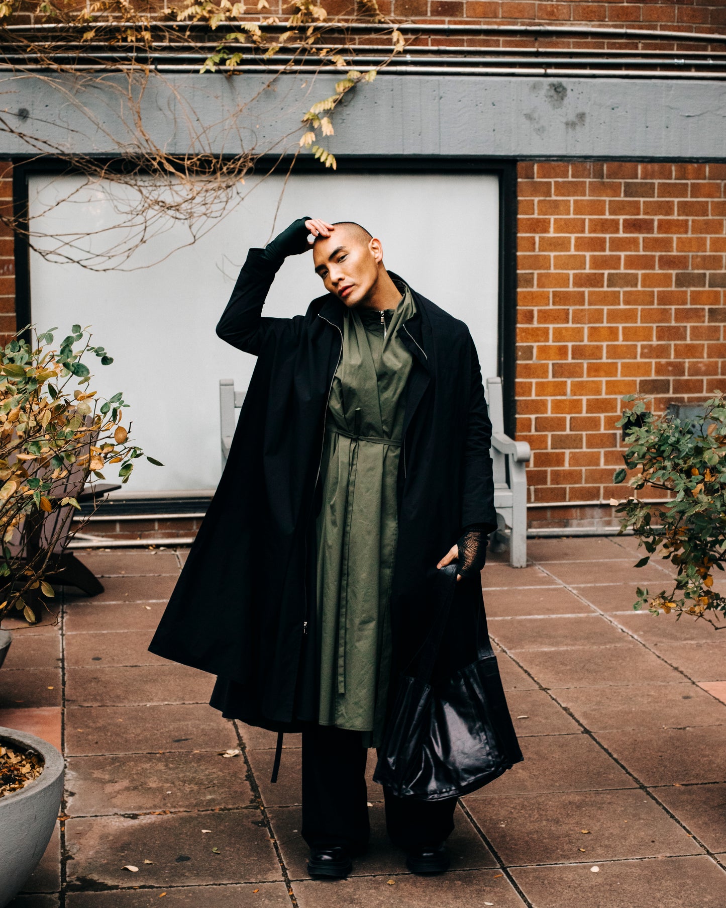 Person in an outdoor urban environment with greenery. Wearing black Japanese nylon raincoat posing with a hand on the head.

Model: @lejamie_