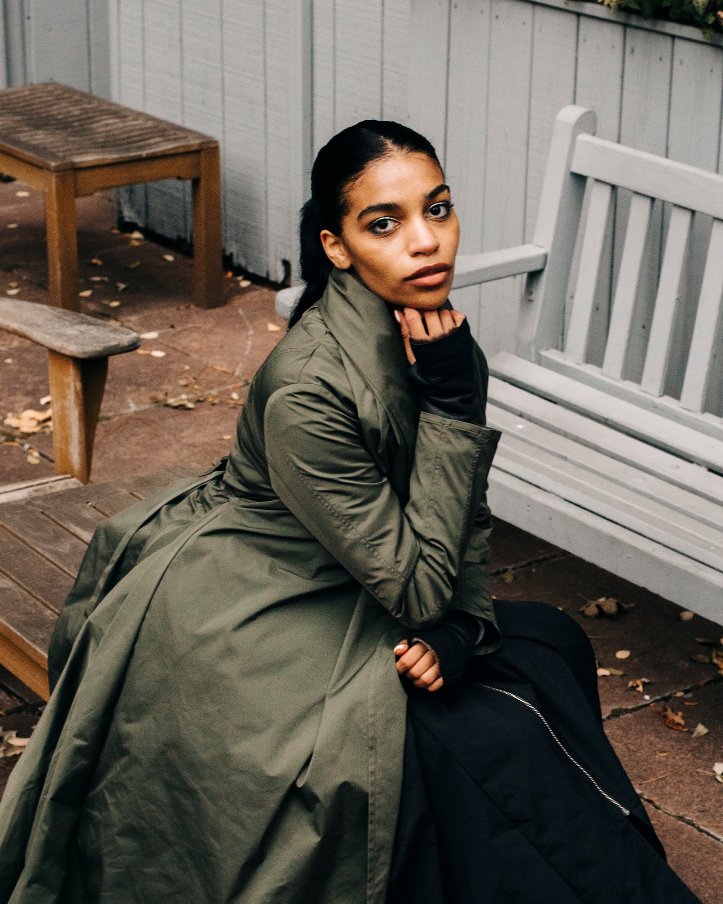 Woman posing on a bench wearing an olive Japanese nylon coat with a hand under her chin.