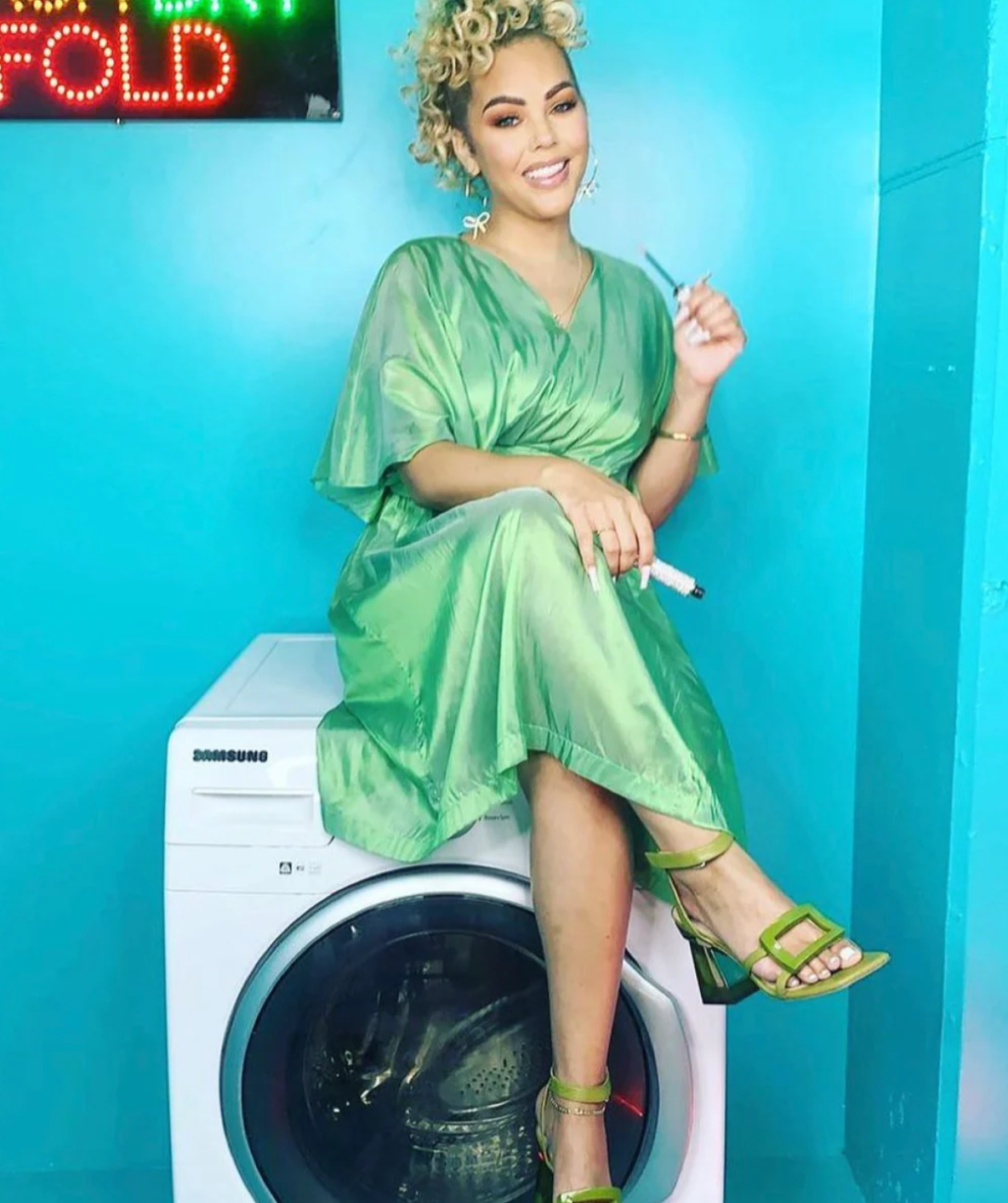 A woman wearing a loose iridescent green dress sitting on top of a washing machine.