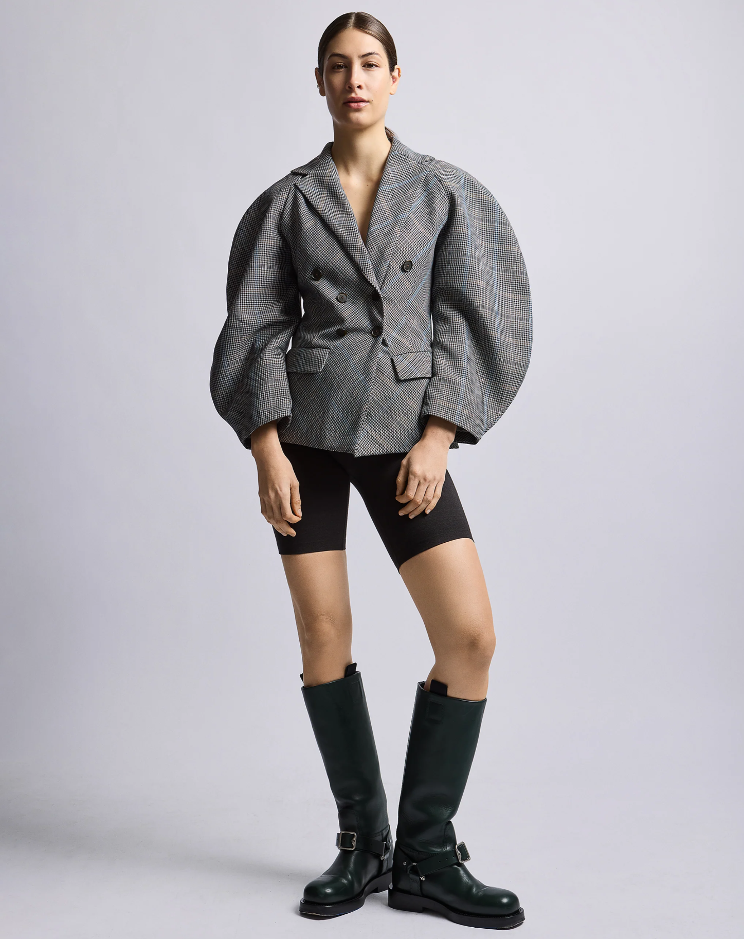Woman standing in a contrapose position wearing a gray double-breasted blazer with plaid patterns.