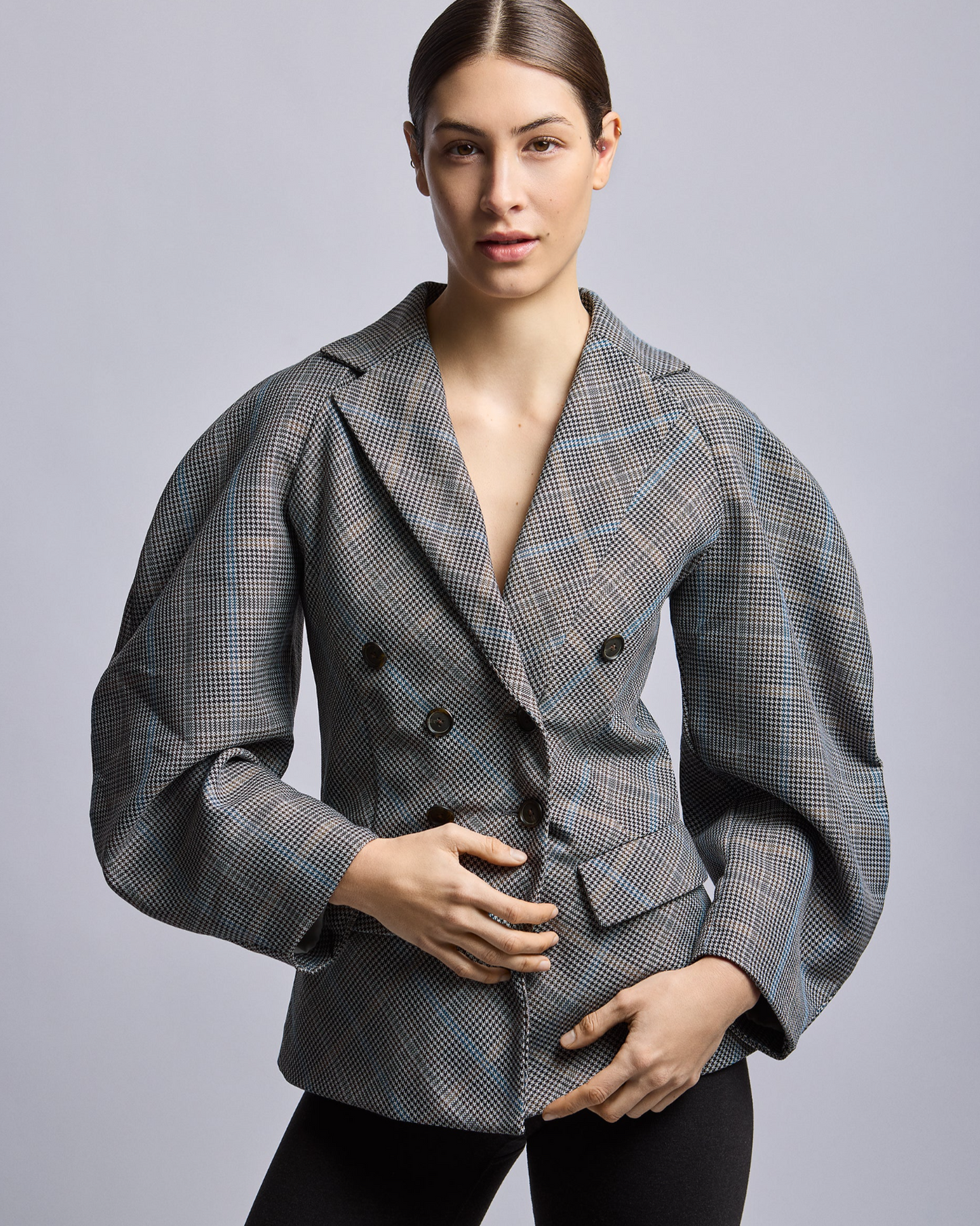 Zoomed in shot of a woman standing in a contrapose position wearing a gray double-breasted blazer with plaid patterns.