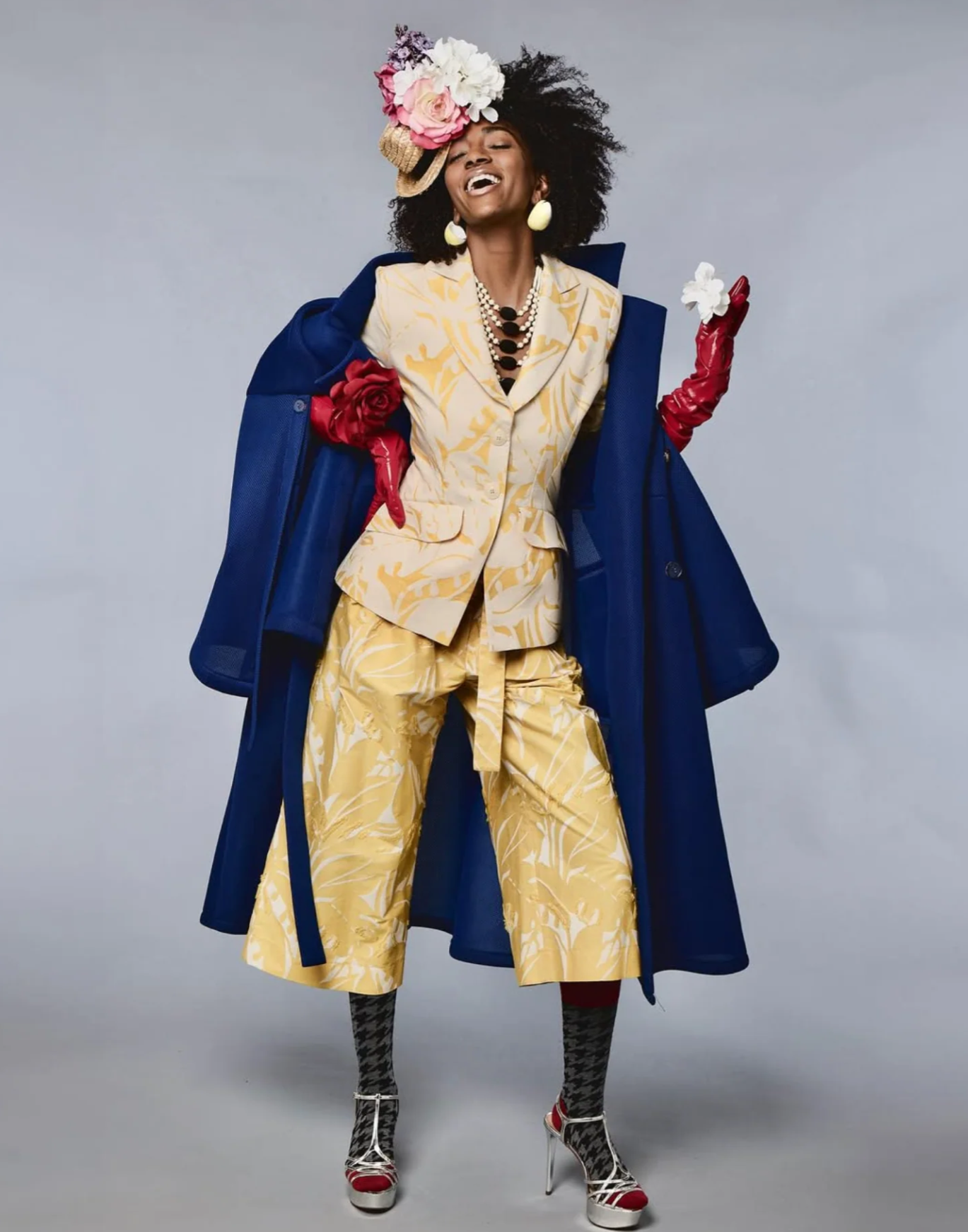 A model posing in a vibrant fashion ensemble featuring a yellow and white patterned suit, a deep blue coat, red gloves, a floral hat, and lace-up heels, set against a plain gray studio background.