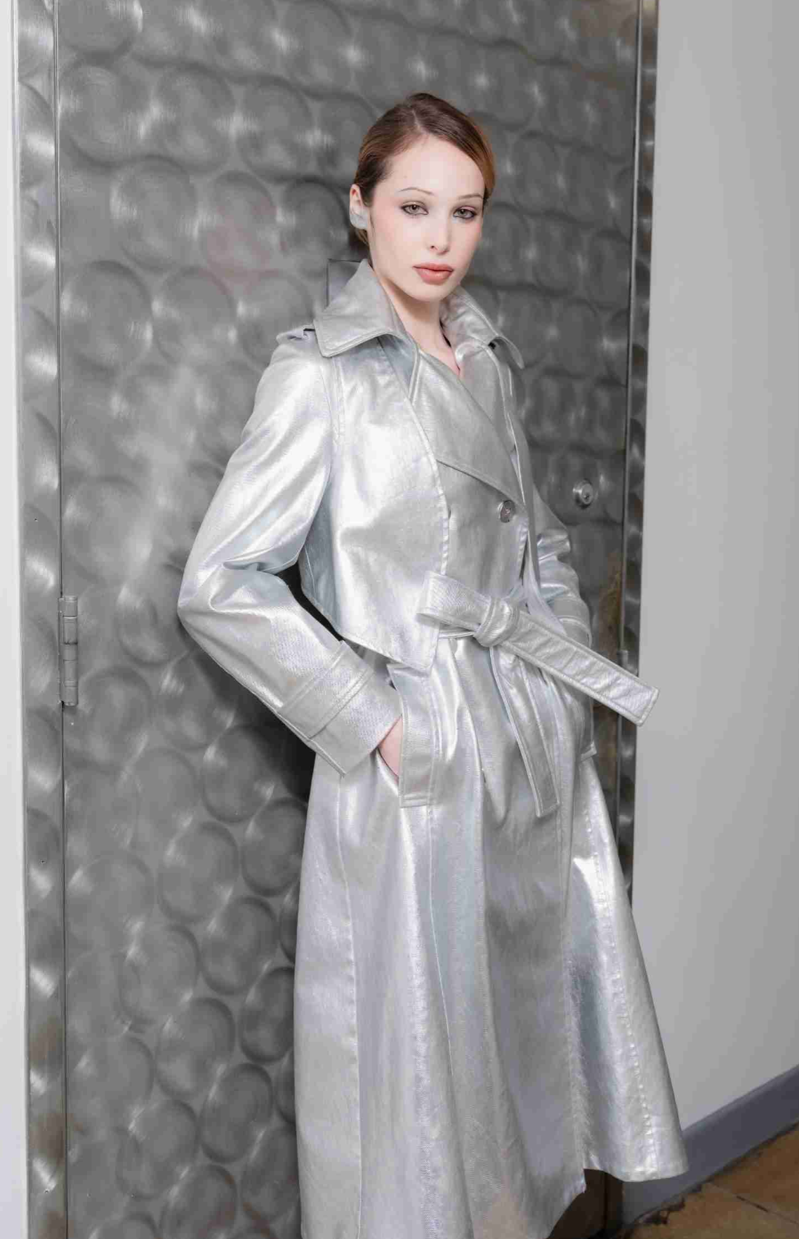 A woman wearing a Long Silver Bristol Trench coat made of satin with a removable vest in textured leather in front of a metallic door.

@natalieturnerr captured beautifully by @_vazhak_ creative eye @kent.nycfashion
@makohairmakenyc
@gabriel.barse
@suecandia
@the_confessional_showroom_ny
@confessional_showroom_miami