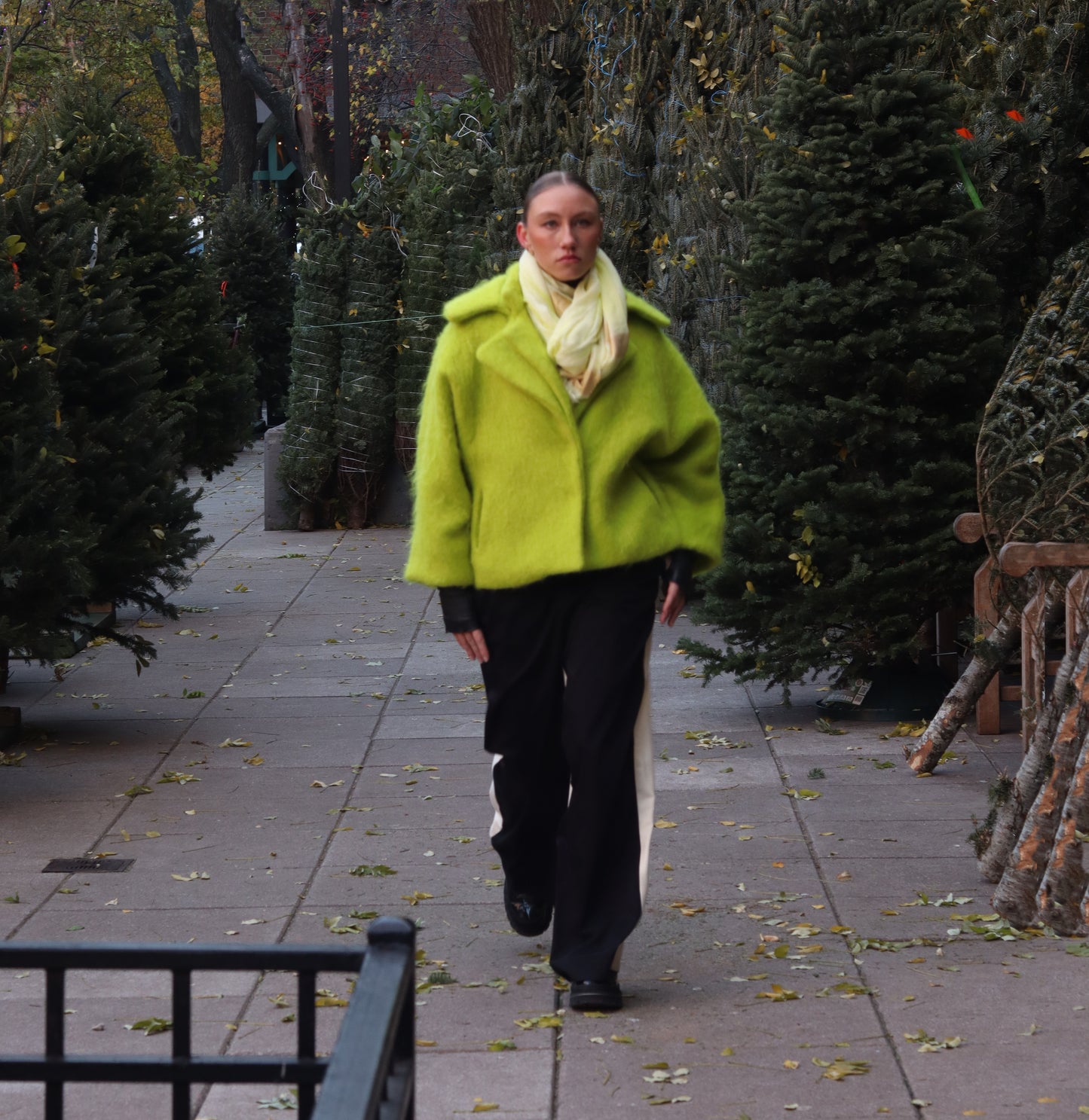 Woman wearing a lime alpaca jacket with a navy dress standing on a sidewalk with evergreen trees in the background.