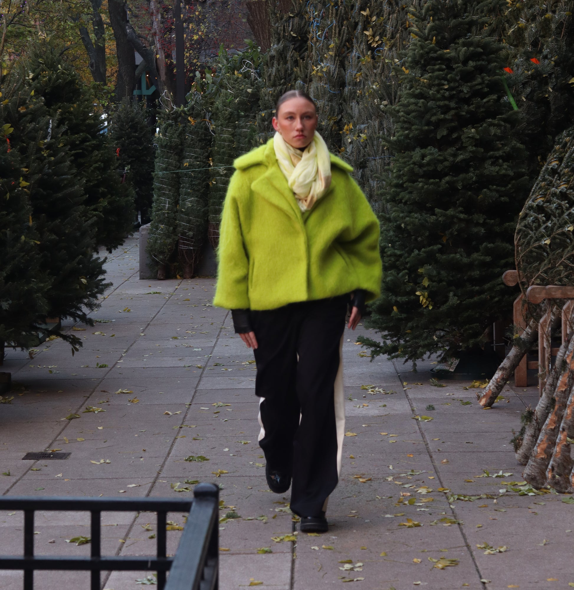 Woman wearing a lime alpaca jacket with a navy dress standing on a sidewalk with evergreen trees in the background.