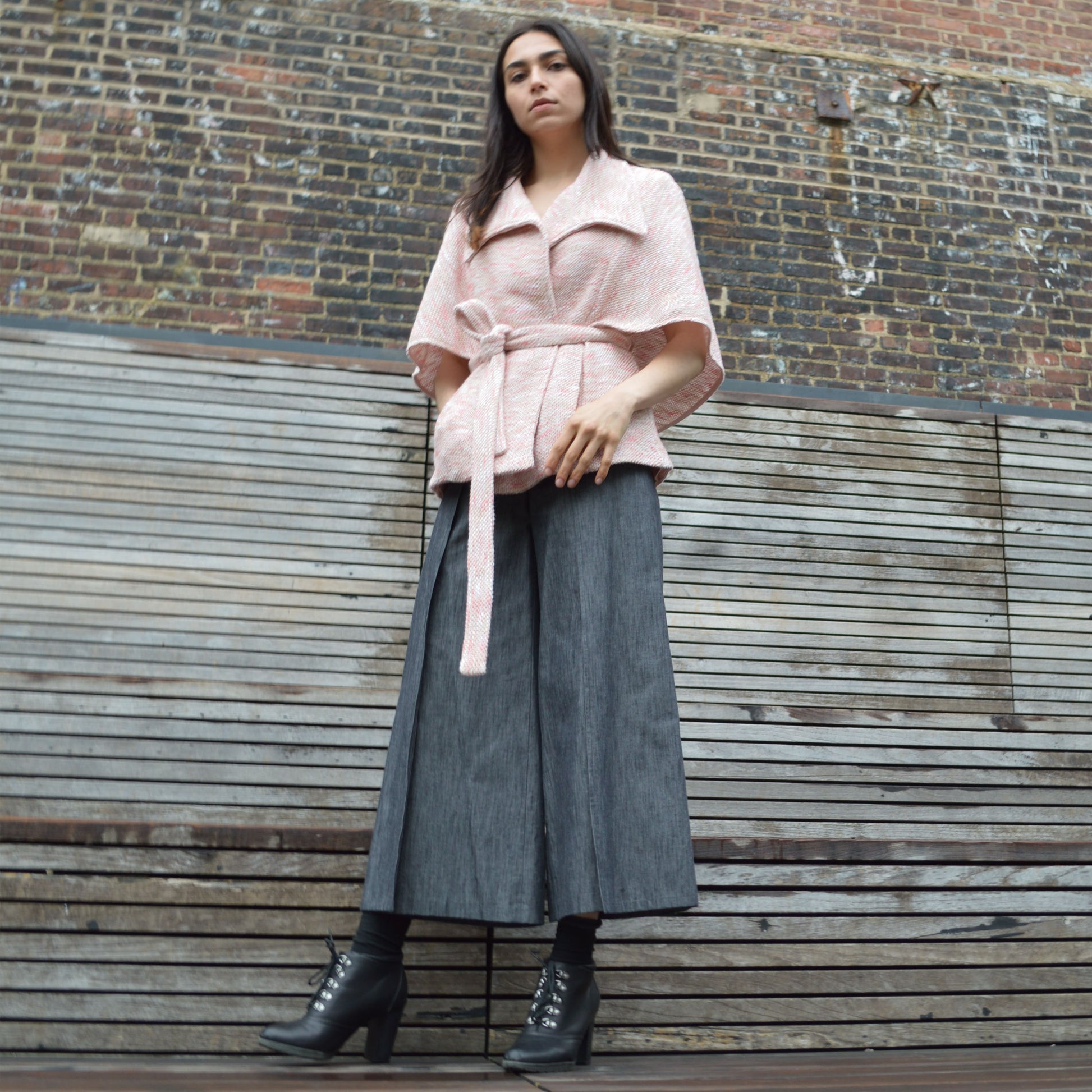 Woman wearing light pink textured wool blend wrap jacket with transformable cape with black fringed detail. Standing in front of  brick and wood plank wall, looking down.