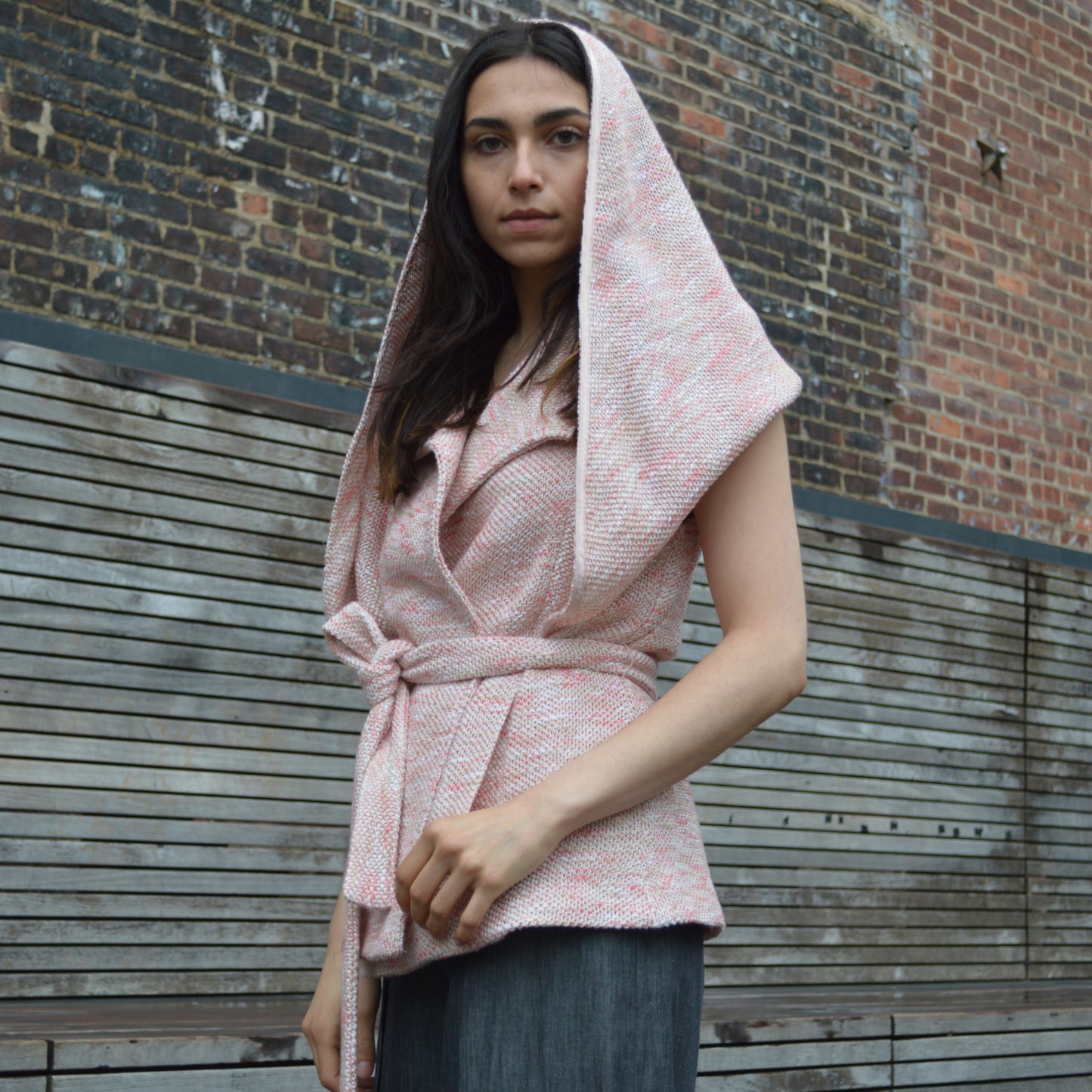 Woman wearing light pink textured wool blend wrap jacket with transformable cape with black fringed detail. Hood is drawn, covering back of head and concealing shoulders. Standing in front of  brick and wood plank wall, looking down.