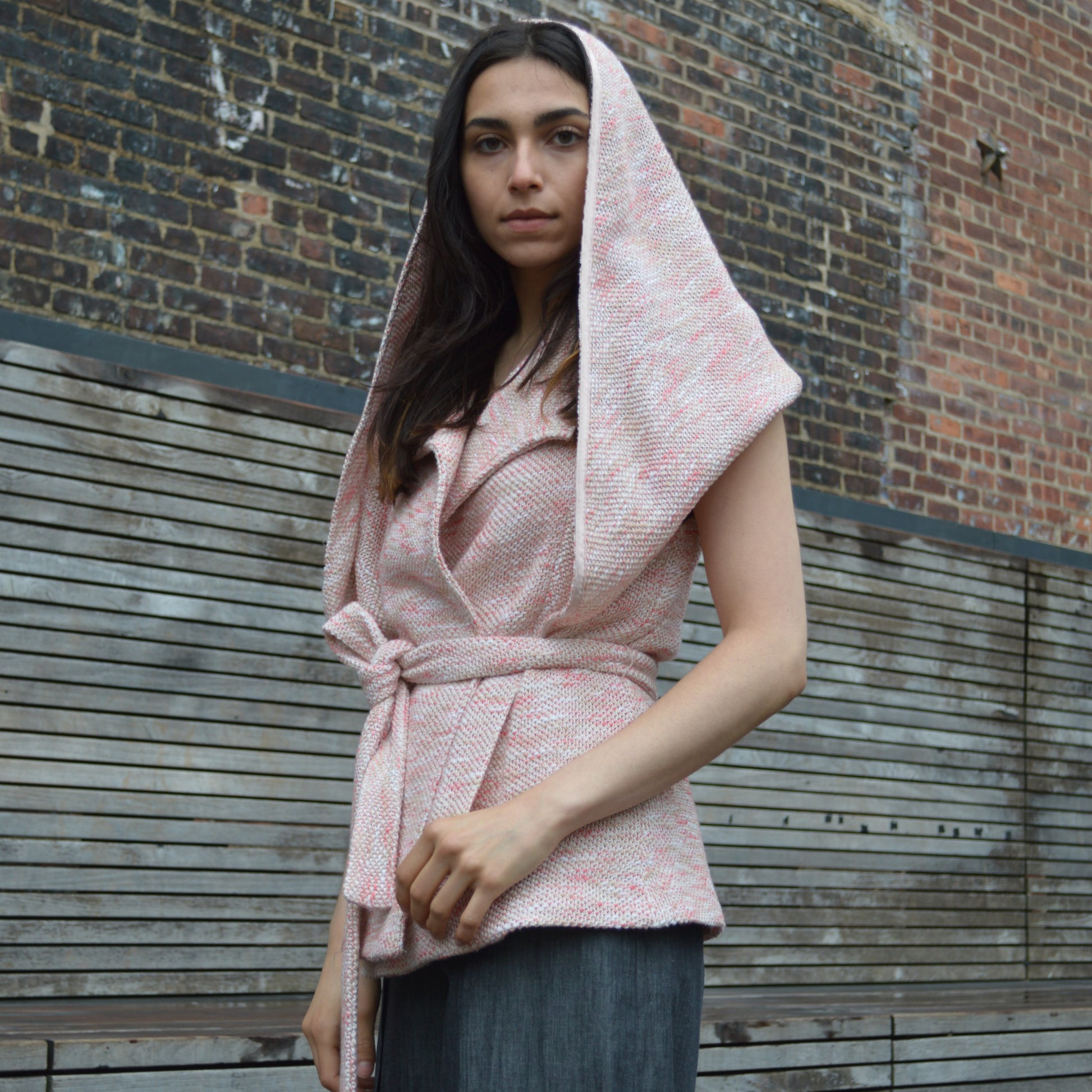 Woman wearing light pink textured wool blend wrap jacket with transformable cape with black fringed detail. Hood is drawn, covering back of head and concealing shoulders. Standing in front of  brick and wood plank wall, looking down.
